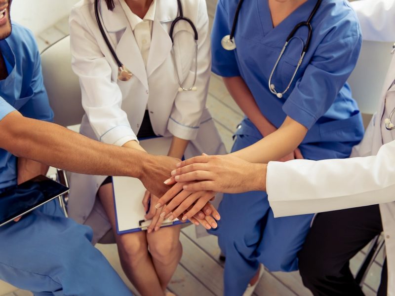 Cropped image of medical doctors of different nationalities and genders holding hands together and smiling, sitting in office